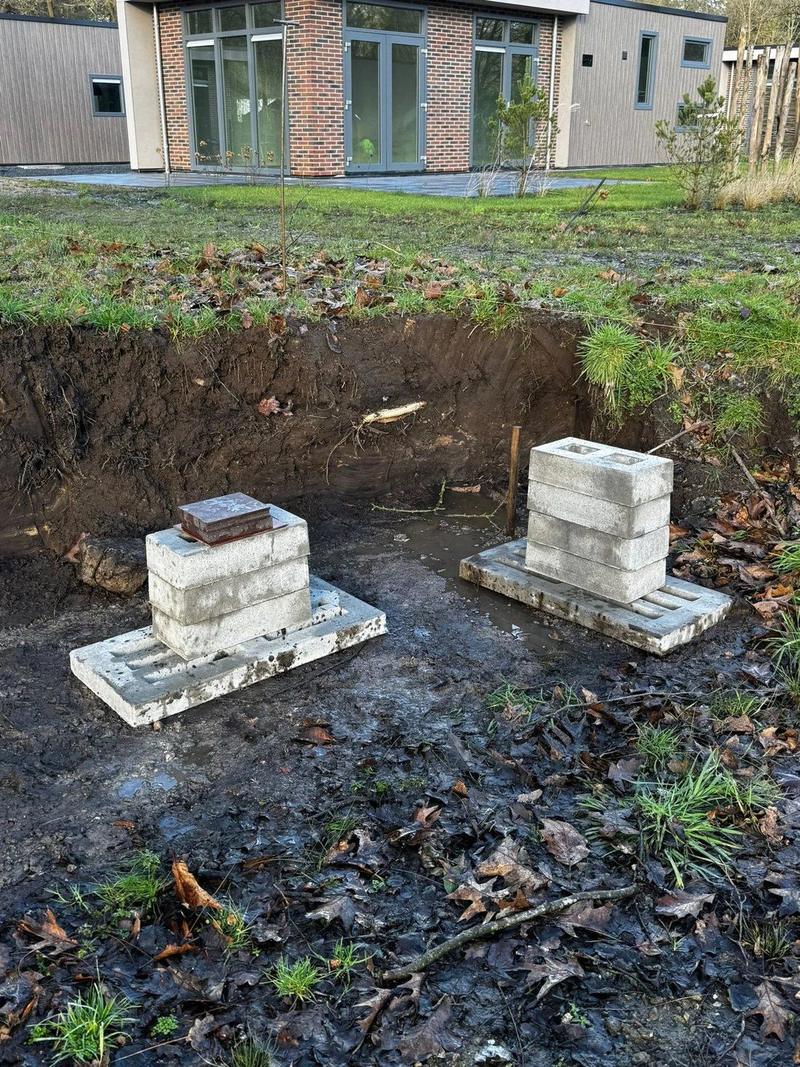 Twee stapels bielzen op grondplaten, met een woning op de achtergrond