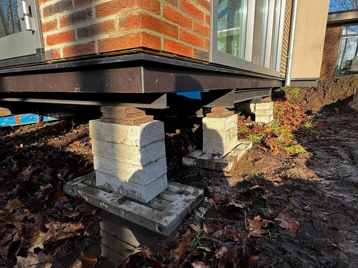 Twee stapels betonnen bielzen op grondplaten dragen de stalen onderbouw van een woning met bakstenen gevel